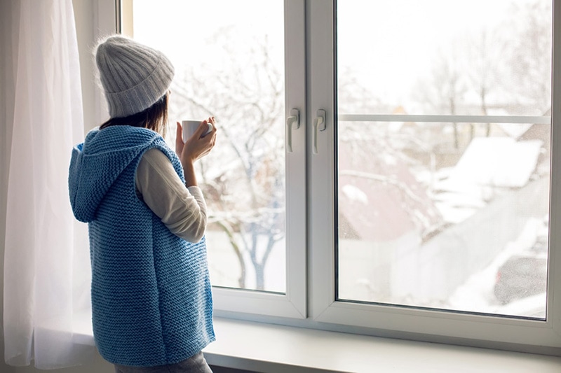 Woman inside wearing a hat and blue vest drinking coffee looking out a window, Why Is My Furnace Blowing Cold Air? | Maintenenace | Wrightsville, PA