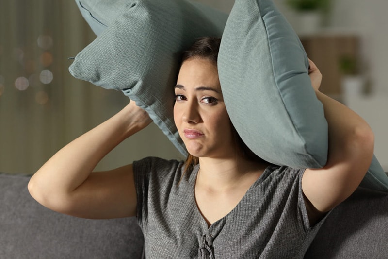 Young brunette woman covering her ears with a pillow, Why is My Air Conditioner Making Noise? | Comfortable Design | HVAC